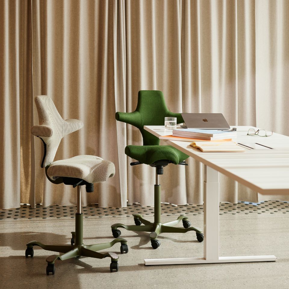 Twee HÅG Capisco bureaustoelen aan een bureau, één in groen en één in een neutrale kleur, veelzijdig inzetbaar in diverse werkomgevingen