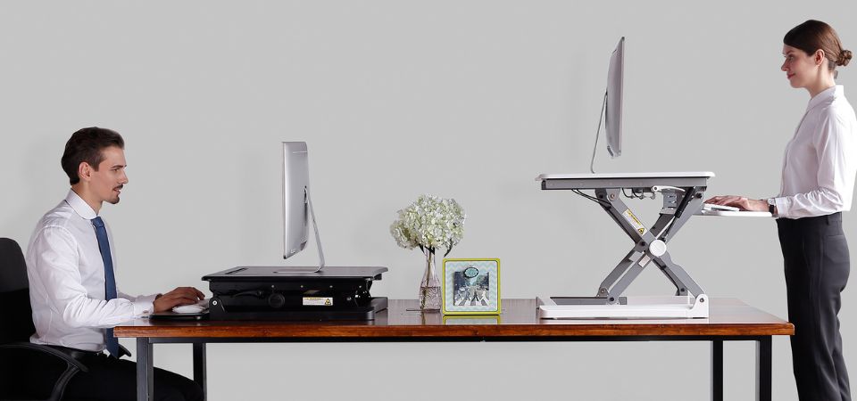 Two people working with the Adjustable Sit-Stand Desk Riser 2: one sitting on the left and one standing on the right, illustrating the sit-stand concept.